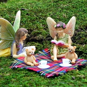 May include: Three fairies sitting on a plaid blanket with two teddy bears and two teacups. One fairy is reading a book, another is holding a book, and the third fairy is looking at the book being read.