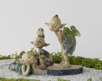 May include: A whimsical fairy garden scene featuring three elf figurines. One elf reclines, another sits with an owl perched on its head, and a third stands with a green dragon on its back. The scene is set on a bed of white pebbles and green moss.
