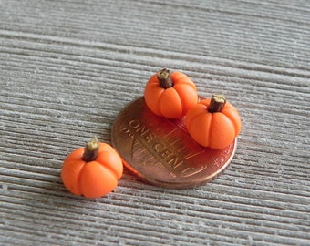 May include: Three miniature orange pumpkins with brown stems rest on a penny. The pumpkins are small, with ribbed sides, and are likely handmade. The penny shows the text "ONE CENT".
