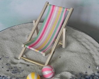 May include: A miniature beach scene with a white wooden beach chair with a pink, yellow, and green striped fabric. Two small beach balls, one pink and white and one yellow and white, are on the sand.