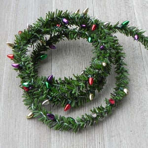 May include: A green garland with colorful Christmas lights. The garland is made of artificial pine needles and features small, teardrop-shaped lights in red, gold, purple, and green. The lights are evenly spaced along the garland.