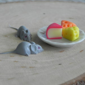 May include: Two gray toy mice looking at a plate of three different colored cheese wedges. The cheese wedges are pink, orange, and yellow.
