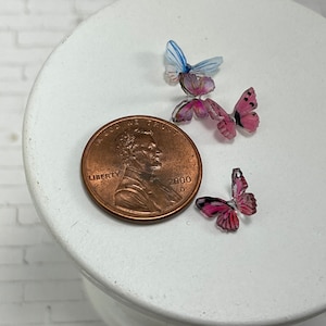 May include: Three small butterfly decorations, two pink and one blue, are arranged on a white surface next to a US penny dated 2000.