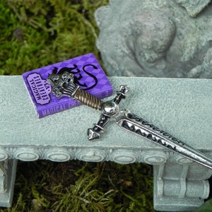 May include: A miniature silver sword with a black and silver handle lies on a grey stone bench next to a small purple book titled "Magical Creatures Vol. 3".