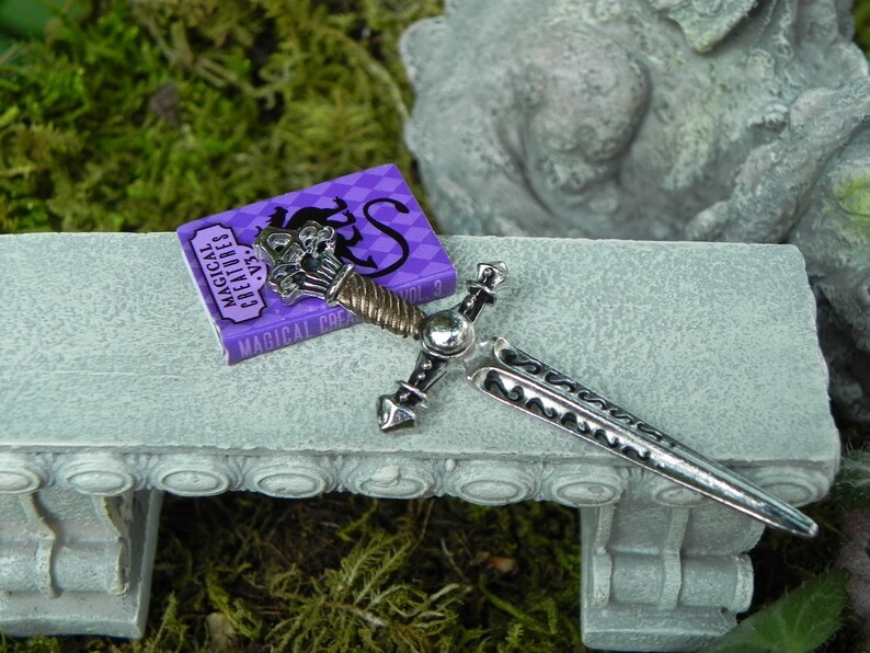 May include: A miniature silver sword with a black and silver handle lies on a grey stone bench next to a small purple book titled "Magical Creatures Vol. 3".
