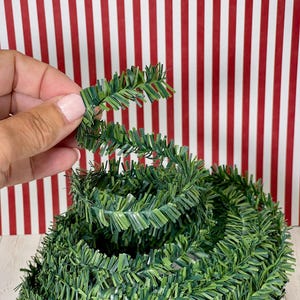 May include: A roll of green artificial garland with a detailed, textured design. The garland is held by a hand against a red and white striped background. The garland is a festive decoration.