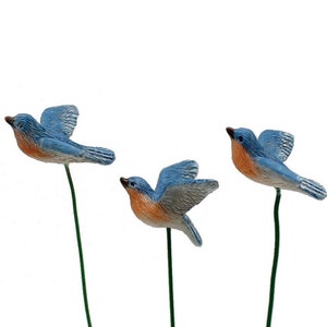 May include: Three bluebird figurines with outstretched wings, perched on green stems. The birds have blue wings and backs, with orange chests and brown beaks. The background is plain white.