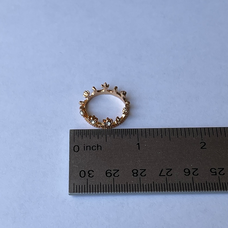 May include: A gold-toned ring with a crown design and clear rhinestones. The ring is measured against a ruler, showing it is less than 1 inch in diameter.