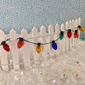 May include: A miniature white picket fence with a string of colorful Christmas lights. The lights are red, orange, yellow, green, and blue. The scene is set in a snowy landscape with a light blue background.