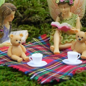 May include: A whimsical scene featuring miniature fairy figurines and teddy bears enjoying a picnic. The scene includes a red, green, and black plaid blanket, white teacups, and a lush green moss background. The fairies are reading books.