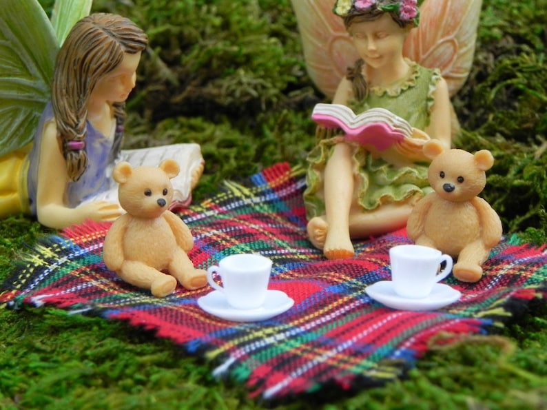 May include: A whimsical scene featuring miniature fairy figurines and teddy bears enjoying a picnic. The scene includes a red, green, and black plaid blanket, white teacups, and a lush green moss background. The fairies are reading books.
