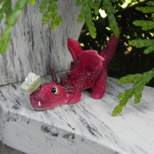 May include: A small, pink, dragon figurine with a butterfly on its head. The dragon has a long tail and is sitting on a white wooden surface.
