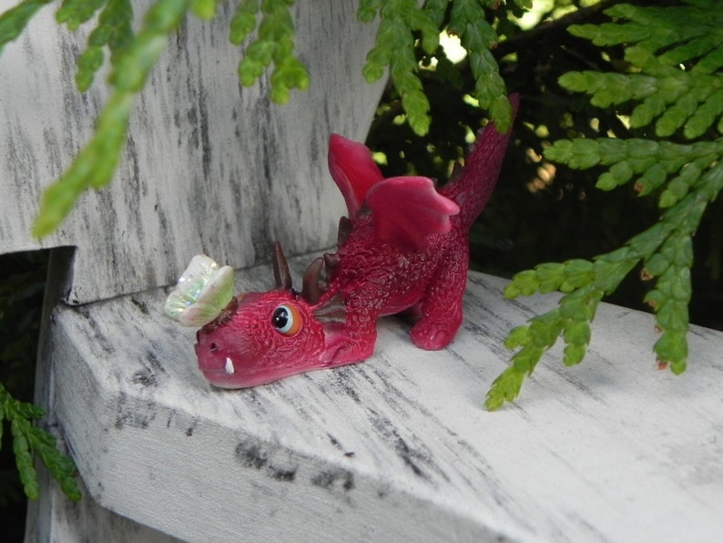 May include: A small, pink, dragon figurine with a butterfly on its head. The dragon has a long tail and is sitting on a white wooden surface.