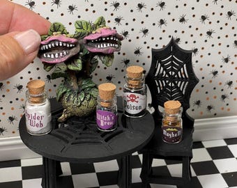 以下が含まれることがあります： 黒いテーブルと椅子を備えた、ハロウィーンをテーマにしたミニチュアシーン。 テーブルには、「Spider Web」、「Witch's Brew」、「Poison」、「Mayhem」とラベル付けされた小さなガラス瓶が置かれています。 ピンク色の歯のような花を持つ植物がテーブルの上に置かれています。