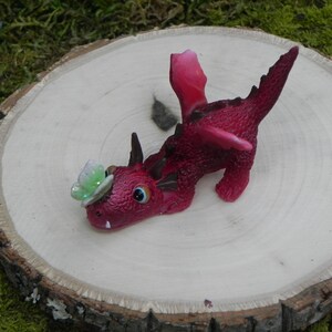 May include: A small, pink dragon figurine with a green butterfly on its head. The dragon is lying on its side with its wings spread out. It is on a wooden surface.