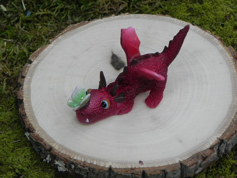 May include: A small, pink dragon figurine with a green butterfly on its head. The dragon is lying on its side with its wings spread out. It is on a wooden surface.