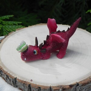 May include: A small, red, plastic dragon figurine with a green butterfly on its head. The dragon is posed in a playful, crouching position with its wings spread out.