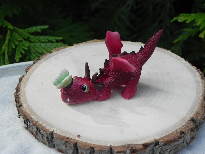 May include: A small, red, plastic dragon figurine with a green butterfly on its head. The dragon is posed in a playful, crouching position with its wings spread out.
