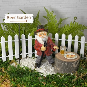 May include: A miniature Santa Claus figurine holding a microphone and standing in front of a white picket fence with a sign that reads "Beer Garden".