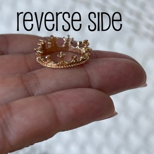May include: Gold crown ring with a detailed design. The ring is on a finger.