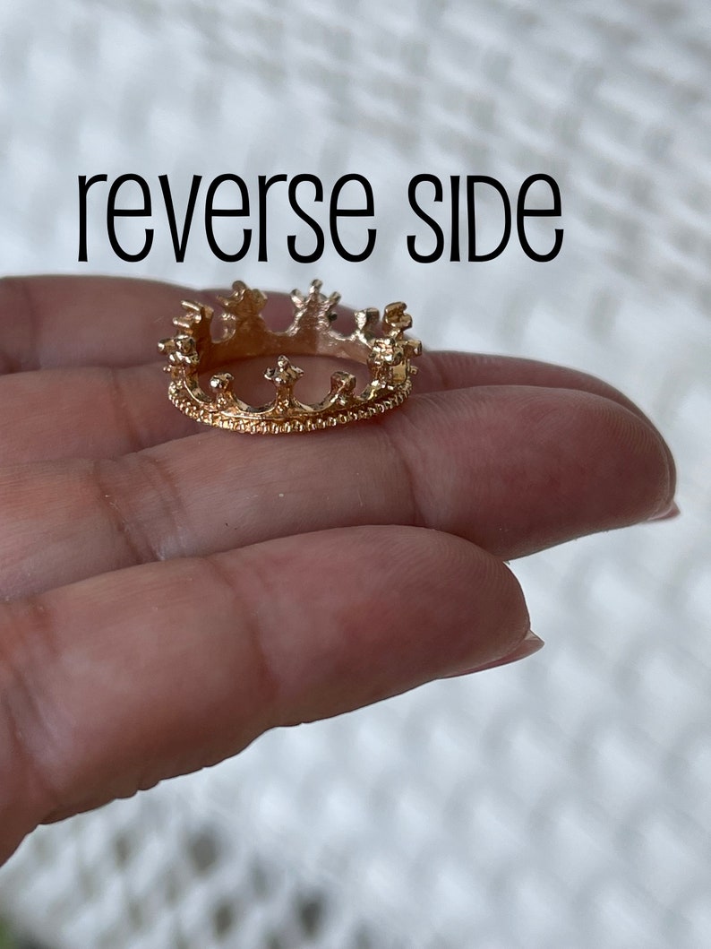 May include: Gold crown ring with a detailed design. The ring is on a finger.