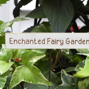 Op de afbeelding: Een rechthoekig houten bord met de tekst "Enchanted Fairy Garden" in bruine letters. Het bord is wit met een verweerde afwerking en staat tussen groen en wit gevlekt klimop.
