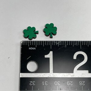 May include: Two green four-leaf clover shapes made of wood, measured against a ruler. The clovers are approximately 1 inch in size.