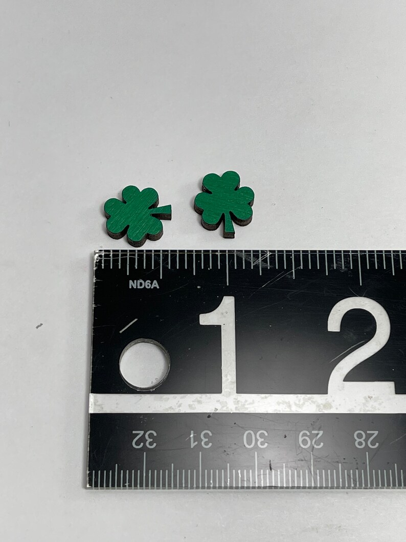 May include: Two green four-leaf clover shapes made of wood, measured against a ruler. The clovers are approximately 1 inch in size.