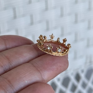 May include: A gold crown ring with small clear stones. The ring is on a finger.