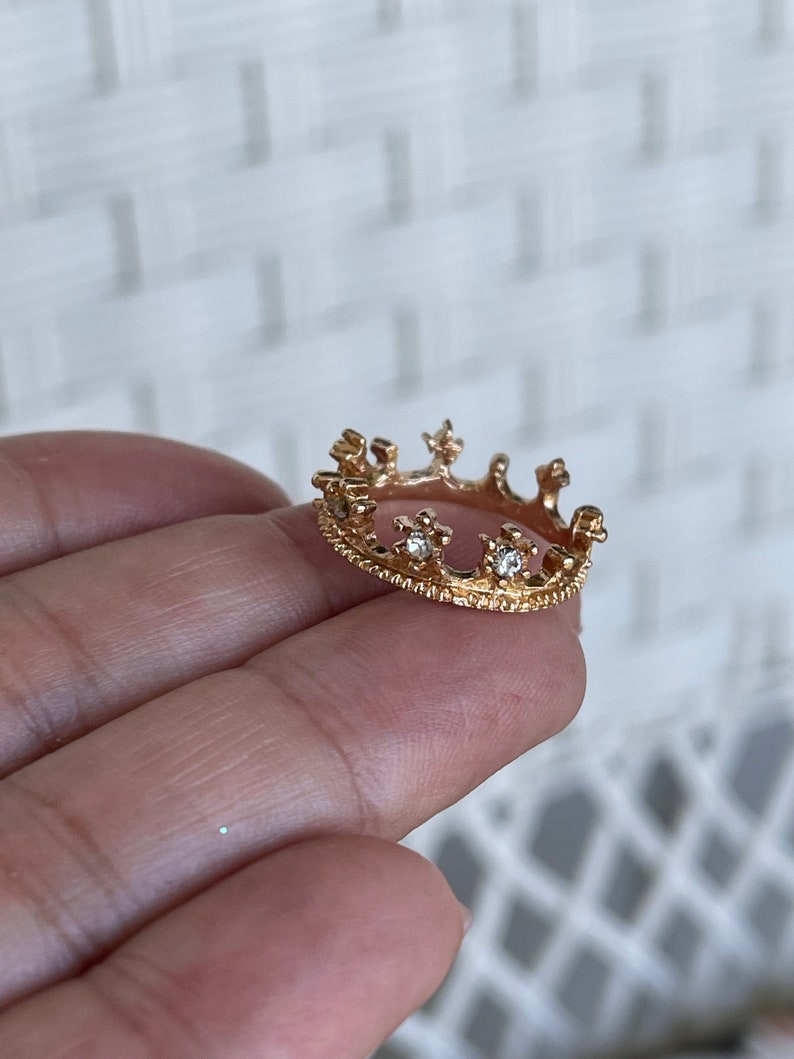May include: A gold crown ring with small clear stones. The ring is on a finger.
