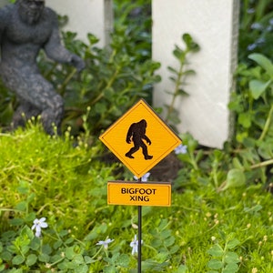 May include: A yellow and black warning sign with a silhouette of a Bigfoot walking. The sign reads "BIGFOOT XING" and is on a black metal stake in a bed of green moss.