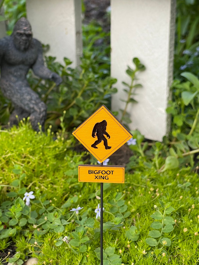 May include: A yellow and black warning sign with a silhouette of a Bigfoot walking. The sign reads "BIGFOOT XING" and is on a black metal stake in a bed of green moss.