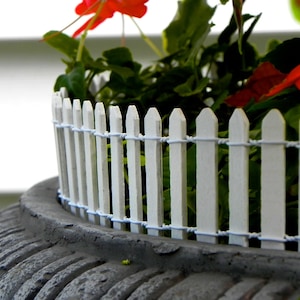 May include: A white picket fence surrounds a potted plant with red flowers. The pot is made of a dark gray material and has a textured surface.