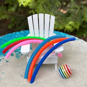 May include: A miniature white Adirondack chair with five colourful pool noodles draped over the back and arms. A miniature rainbow striped beach ball sits on the white sand below the chair.