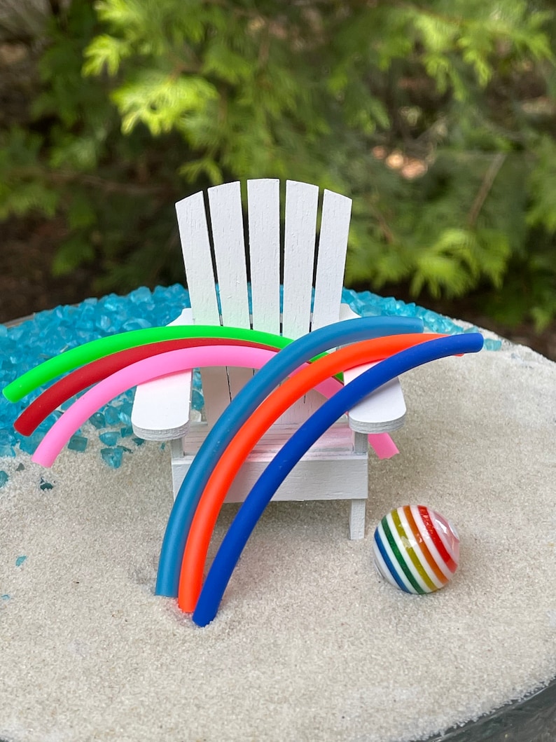 May include: A miniature white Adirondack chair with five colourful pool noodles draped over the back and arms. A miniature rainbow striped beach ball sits on the white sand below the chair.
