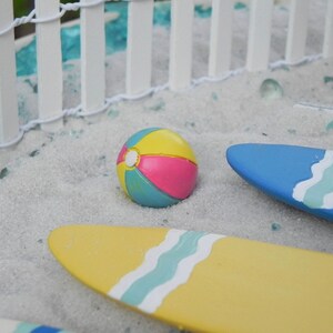 May include: A miniature beach scene with a white picket fence, blue and yellow surfboards, and a colorful beach ball.