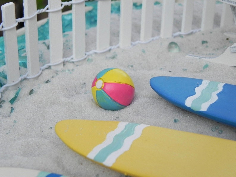 May include: A miniature beach scene with a white picket fence, blue and yellow surfboards, and a colorful beach ball.