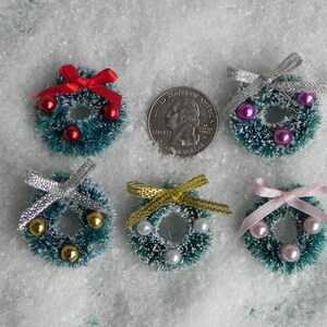 May include: Six miniature Christmas wreaths with different colored bows and ornaments. The wreaths are green with a white, fluffy texture. The ornaments are red, pink, gold, and silver.