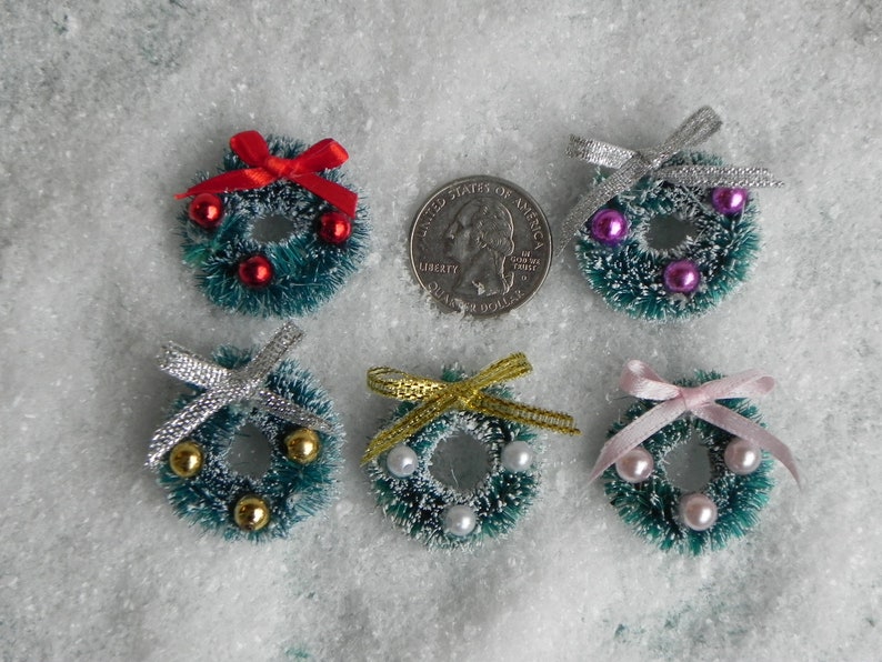 May include: Six miniature Christmas wreaths with different colored bows and ornaments. The wreaths are green with a white, fluffy texture. The ornaments are red, pink, gold, and silver.