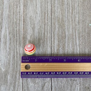 May include: A small, round, colourful ceramic knob with a white centre and red, orange, yellow, and green stripes. The knob is approximately 2.5 cm in diameter and is resting on a wooden surface.