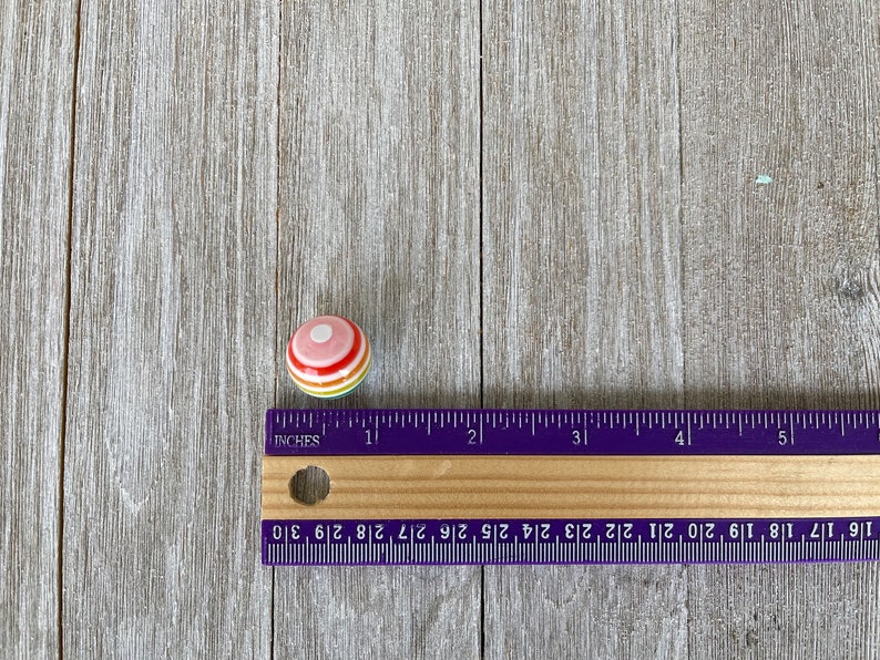 May include: A small, round, colourful ceramic knob with a white centre and red, orange, yellow, and green stripes. The knob is approximately 2.5 cm in diameter and is resting on a wooden surface.