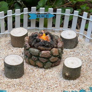K&ouml;nnte beinhalten: Ein Miniatur-Steinkamin mit einer gl&uuml;henden orangefarbenen Flamme, umgeben von vier Holzst&uuml;mpfen. Der Kamin befindet sich auf einer wei&szlig;en Sandfl&auml;che mit blauen Glassteinen am Rand. Ein wei&szlig;er Lattenzaun mit einem blauen Schild, auf dem "BEACH" steht, befindet sich im Hintergrund.