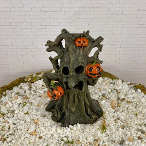 May include: A spooky Halloween tree figurine with a carved face and three orange jack-o'-lanterns. The tree is dark brown with a textured bark effect. The pumpkins have carved faces and are orange. The figurine is set on a bed of white pebbles.