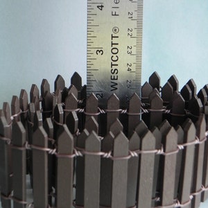 May include: A brown wooden picket fence with wire connecting the pickets. The fence is approximately 2 inches tall and is curled into a circle. A ruler is in the background to show the size of the fence.