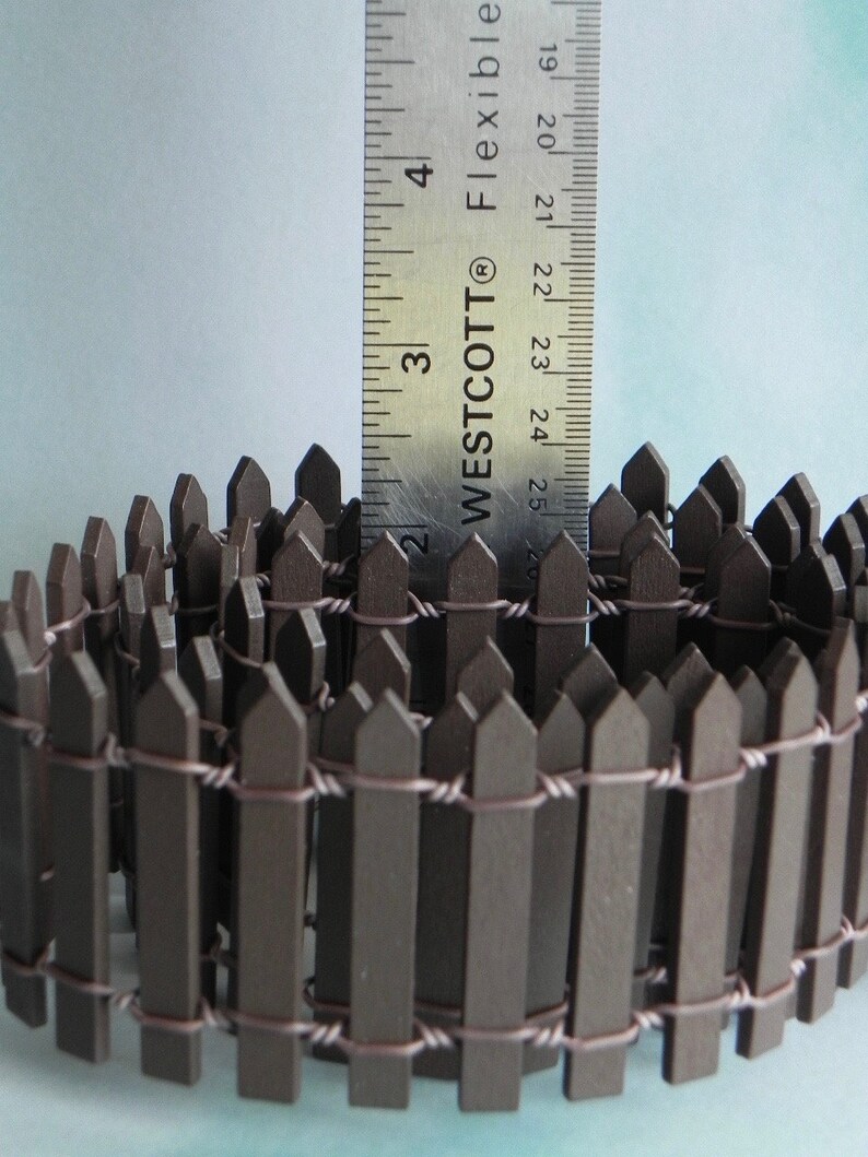 May include: A brown wooden picket fence with wire connecting the pickets. The fence is approximately 2 inches tall and is curled into a circle. A ruler is in the background to show the size of the fence.