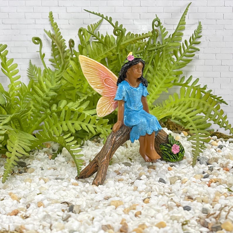 May include: A miniature fairy figurine with a blue dress and peach-colored wings sits on a brown branch. The fairy has black hair and a pink flower in her hair. Green ferns and white pebbles surround the fairy.