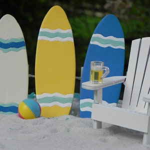May include: A miniature beach scene with three surfboards, a white Adirondack chair, a small glass of beer, and a beach ball. The surfboards are painted in yellow, white, and blue with wavy lines. The chair has a small table attached to the armrest. The beach ball is yellow, pink, and blue.