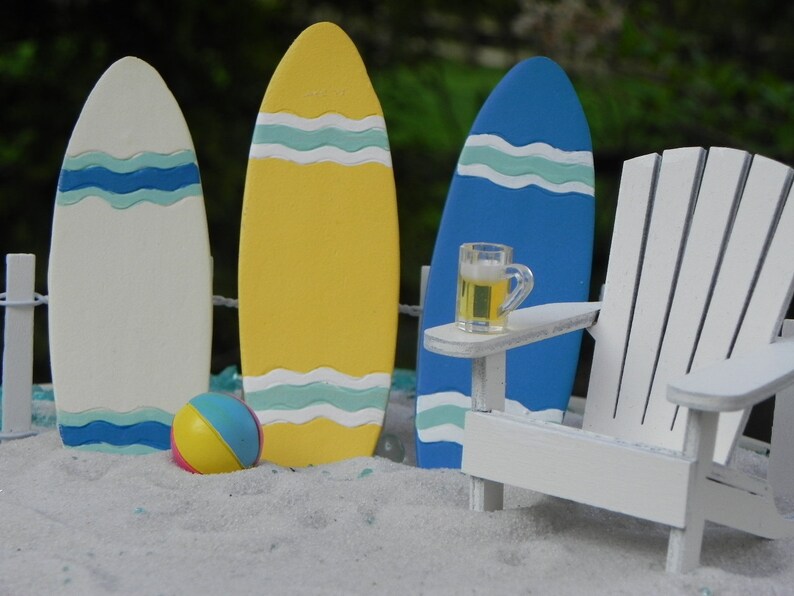 May include: A miniature beach scene with three surfboards, a white Adirondack chair, a small glass of beer, and a beach ball. The surfboards are painted in yellow, white, and blue with wavy lines. The chair has a small table attached to the armrest. The beach ball is yellow, pink, and blue.