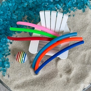 May include: A white miniature beach chair with colourful rubber pool noodles and a striped beach ball on a bed of blue and white sand.