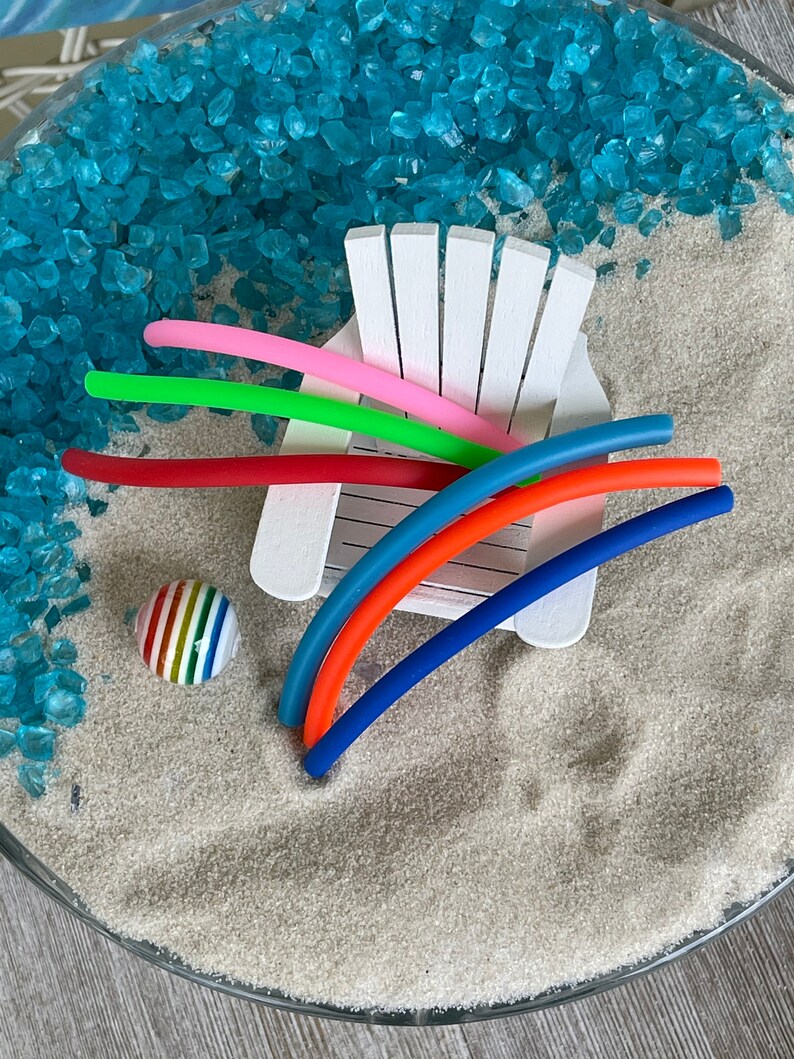 May include: A white miniature beach chair with colourful rubber pool noodles and a striped beach ball on a bed of blue and white sand.